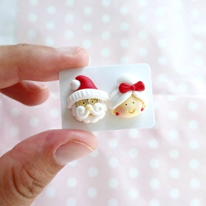 May include: A pair of white polymer clay earrings featuring Santa Claus and Mrs. Claus. The earrings are on a white card.
