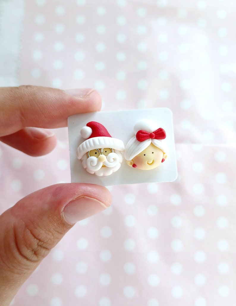 May include: A pair of white polymer clay earrings featuring Santa Claus and Mrs. Claus. The earrings are on a white card.