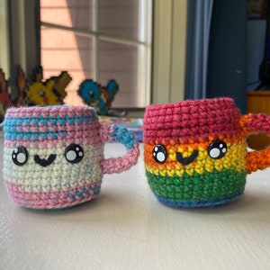 May include: Two crocheted mugs with smiling faces. One mug is pink, blue and white, and the other is red, orange, yellow, green and blue.