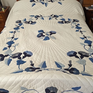 May include: A white quilt with a blue and white floral pattern. The quilt is made with a variety of fabrics and is quilted in a traditional style. The quilt is displayed on a bed with a wooden headboard.