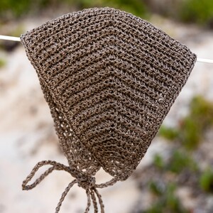 Puede incluir: Un sombrero de sol marrón tejido a ganchillo con una corona puntiaguda y un cierre de lazo. El sombrero está hecho de un material tejido texturizado y está suspendido contra un fondo borroso de vegetación y arena.