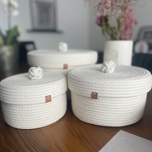 Peut inclure: Trois paniers de rangement en corde blanche avec couvercles et accents en cuir marron. Les paniers sont ronds et ont une texture tissée.
