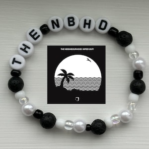 Pode incluir: Uma pulseira de contas com as letras "THE NBHD" em contas brancas. A pulseira inclui contas de lava pretas, contas de pérolas brancas e um pequeno gráfico a preto e branco de uma palmeira e ondas. O texto "THE NEIGHBOURHOOD WIPED OUT!" também é visível.