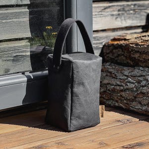 May include: A rectangular, dark gray doorstop with a textured, matte finish. It features a sturdy, matching dark gray fabric handle attached to the top with two metal rivets. A small, light-colored rectangular tag is visible on the side.