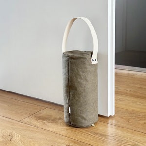 May include: A cylindrical olive green doorstop with a textured fabric finish and a sturdy off-white handle attached with metal rivets. The object stands upright, featuring a vertical seam along the side and a small white tag near the base.