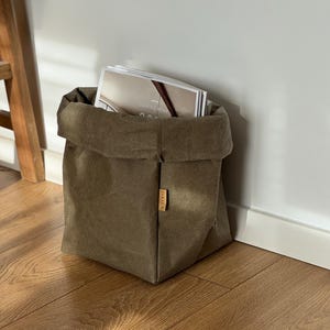 Könnte beinhalten: Eine gefaltete Aufbewahrungstasche aus olivgrünem Canvas mit einem kleinen Holzanhänger. Die Tasche ist mit Zeitschriften gefüllt, auf denen der Text "COMING HOME" zu sehen ist. Die Tasche steht auf einem Holzboden neben einer weißen Wand.