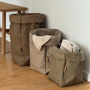 May include: Three upright storage bags in varying shades of brown, tan, and olive green. Each bag features a folded top edge, a square base, and a small rectangular tag on the side. One bag contains a light beige cloth and another holds a magazine.