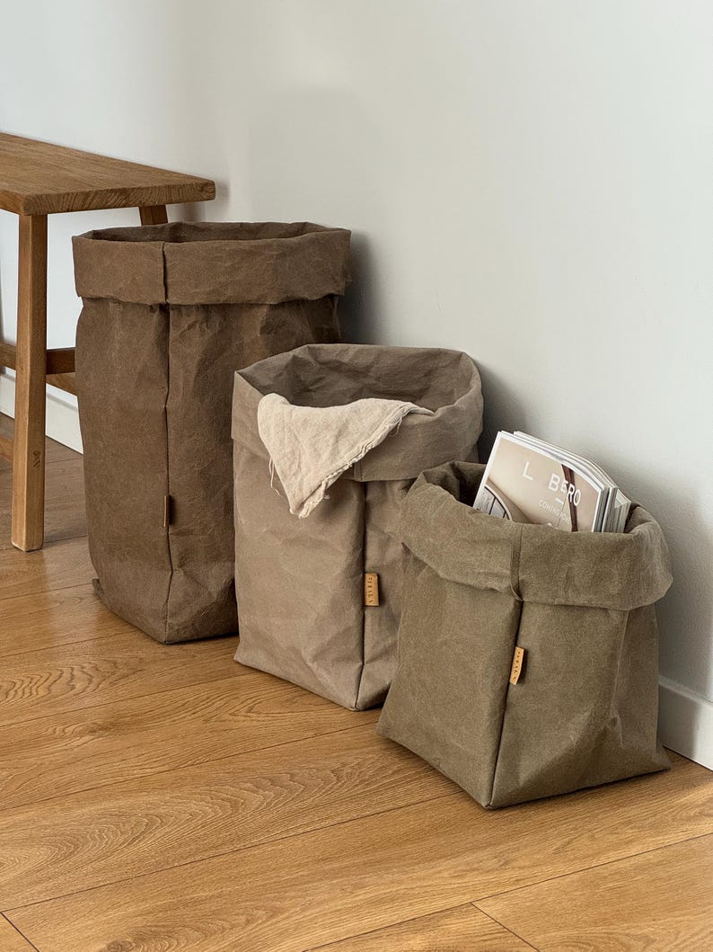 May include: Three upright storage bags in varying shades of brown, tan, and olive green. Each bag features a folded top edge, a square base, and a small rectangular tag on the side. One bag contains a light beige cloth and another holds a magazine.