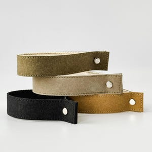 Puede incluir: Cuatro correas de muñeca de ante apiladas en verde oliva, beige, negro y amarillo mostaza. Cada correa tiene un cierre de botón a presión plateado. Las correas son probablemente para una bolsa u otro accesorio.