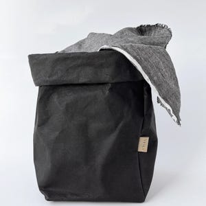 May include: A black, foldable storage bag with a textured, paper-like appearance and a rolled-down top edge. A grey, woven fabric cloth with a frayed edge is draped over the side. A small, rectangular tan label is attached to the side of the bag.