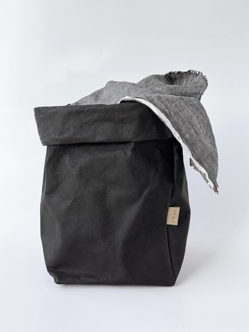 May include: A black, foldable storage bag with a textured, paper-like appearance and a rolled-down top edge. A grey, woven fabric cloth with a frayed edge is draped over the side. A small, rectangular tan label is attached to the side of the bag.
