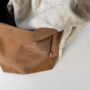 May include: A brown, flexible, paper-like storage basket with a folded top edge and a small side tag, containing a draped, light beige, textured fabric cloth that spills over the rim of the container, creating a soft, layered appearance.
