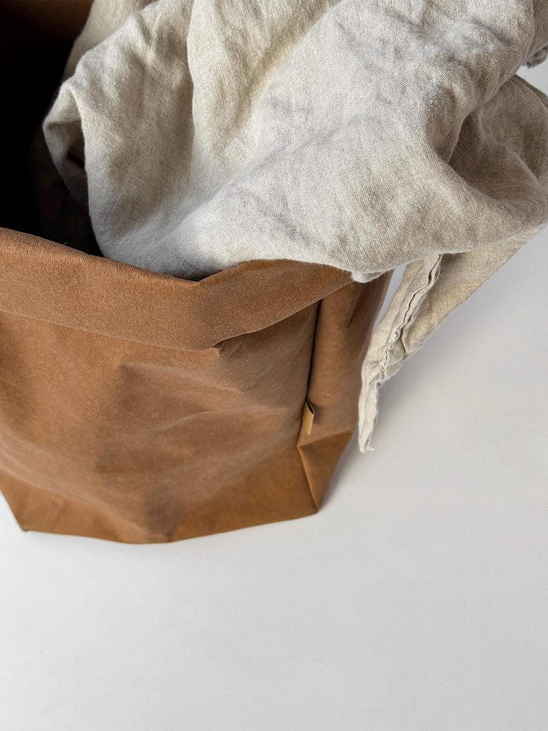 May include: A brown, flexible, paper-like storage basket with a folded top edge and a small side tag, containing a draped, light beige, textured fabric cloth that spills over the rim of the container, creating a soft, layered appearance.