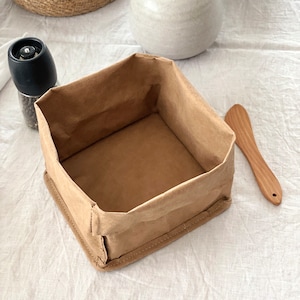 Puede incluir: Una caja de almacenamiento de papel marrón con una parte superior doblada y un borde cosido. La caja está vacía y se encuentra sobre una superficie blanca.