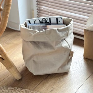 May include: A beige, cylindrical storage bag made of a paper-like material with a folded top edge and a small rectangular tag on the side. The bag is filled with several magazines, including one with the word VOGUE printed in large black letters.
