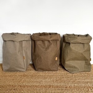 May include: Three upright storage bags in varying shades of brown, tan, and olive green. Each bag features a folded top edge and a small rectangular brand tag attached to the side. The bags have a textured, paper-like appearance with visible creases.