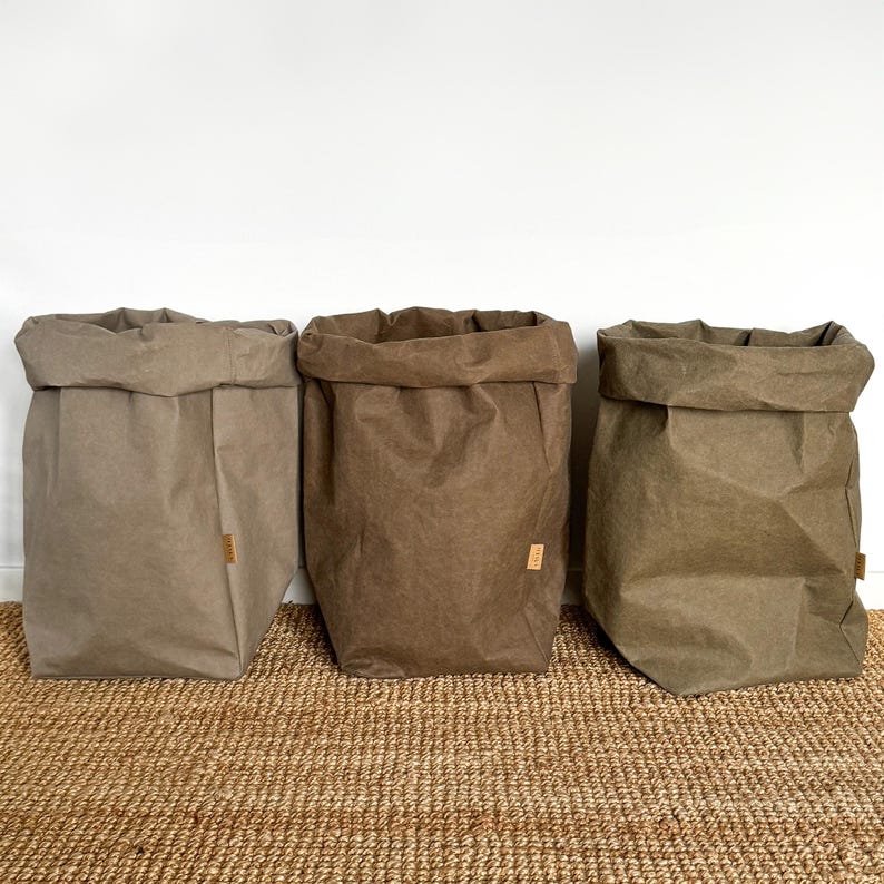 May include: Three upright storage bags in varying shades of brown, tan, and olive green. Each bag features a folded top edge and a small rectangular brand tag attached to the side. The bags have a textured, paper-like appearance with visible creases.
