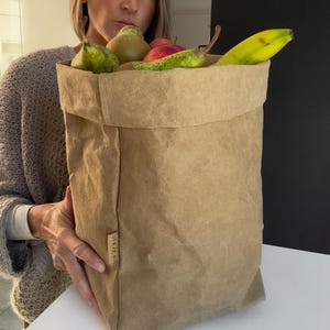 May include: A tan, rectangular, foldable paper storage bag with a textured surface and a small vertical label on the side. The bag is filled with fresh produce, including green pears, a red apple, and a yellow banana, creating a natural display.