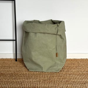 May include: A sage green storage bag made of a textured, paper-like material with a folded top edge. The bag features a rectangular shape with a small, vertical brown fabric label attached to the side seam, displaying subtle, illegible text.