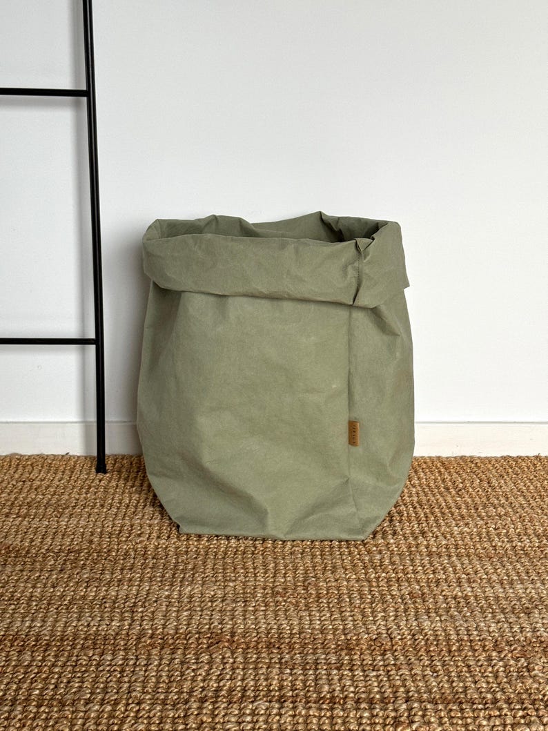 May include: A sage green storage bag made of a textured, paper-like material with a folded top edge. The bag features a rectangular shape with a small, vertical brown fabric label attached to the side seam, displaying subtle, illegible text.