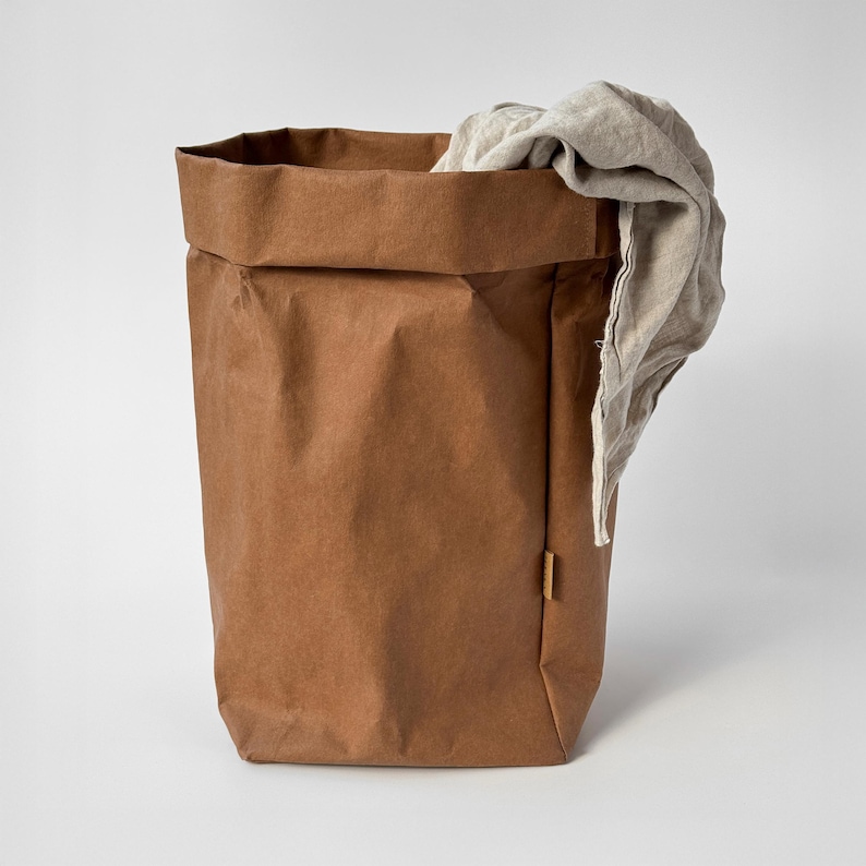 May include: A tall, cylindrical storage bag made of brown, textured material with a folded top edge. A light beige, wrinkled fabric cloth is draped over the right side of the bag, partially hanging inside and over the rim of the container.