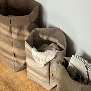 May include: Three cylindrical storage bags made of a textured, paper-like material in varying shades of brown and beige. The bags have folded top edges and small rectangular labels on the side. One bag contains a light-colored cloth and a magazine.