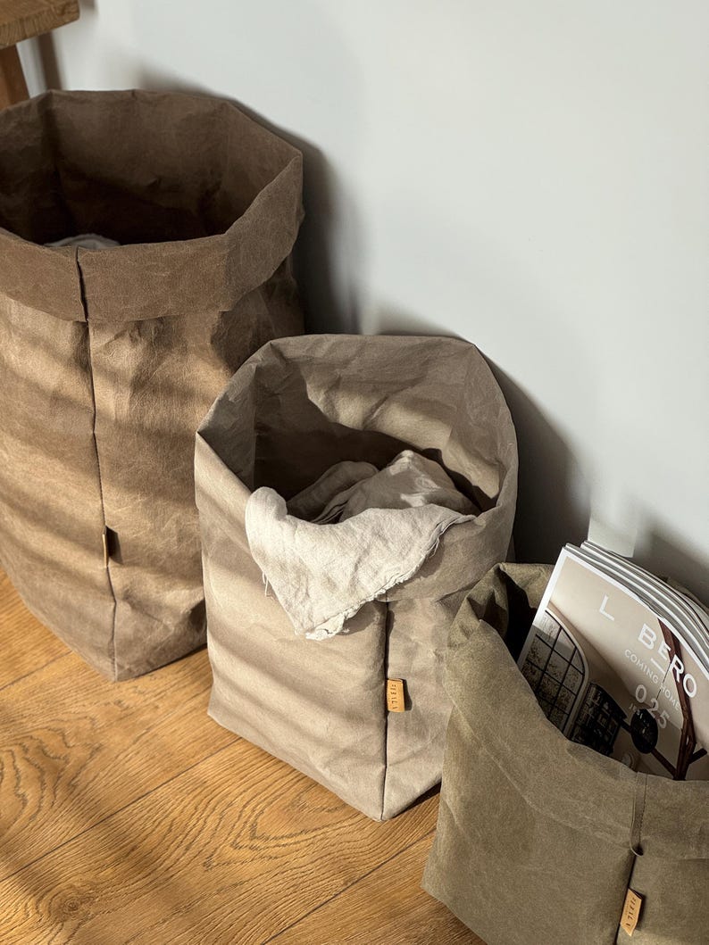 May include: Three cylindrical storage bags made of a textured, paper-like material in varying shades of brown and beige. The bags have folded top edges and small rectangular labels on the side. One bag contains a light-colored cloth and a magazine.