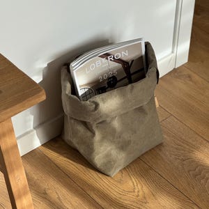 May include: A flexible, olive-green fabric storage bag with a folded top edge, containing a magazine titled LOBERON COMING HOME 2025 JESIEN. The bag has a soft, matte texture and a structured, square-bottomed base, providing a simple storage solution.