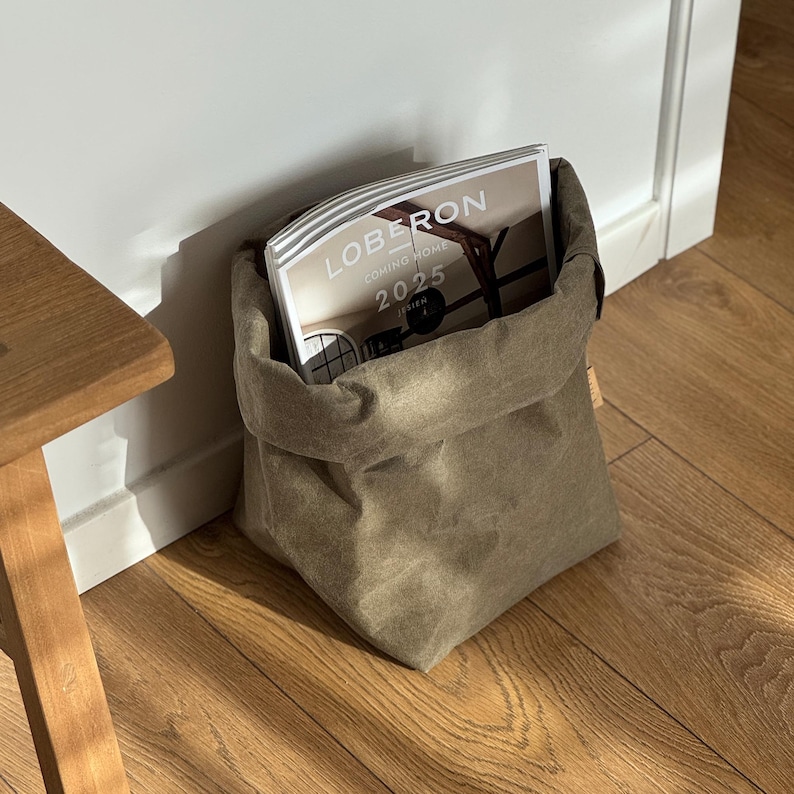 May include: A flexible, olive-green fabric storage bag with a folded top edge, containing a magazine titled LOBERON COMING HOME 2025 JESIEN. The bag has a soft, matte texture and a structured, square-bottomed base, providing a simple storage solution.
