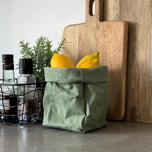 May include: A sage green fabric storage basket with a folded top edge, containing two bright yellow lemons. The basket has a textured, paper-like appearance and a square base, providing a simple container for kitchen items or produce.