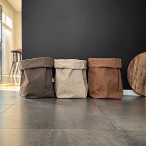 May include: Three rectangular storage bags with folded top edges, arranged in a row. The bags are made of a textured material in shades of dark gray, light beige, and warm tan, each featuring a small rectangular tag on the side seam.