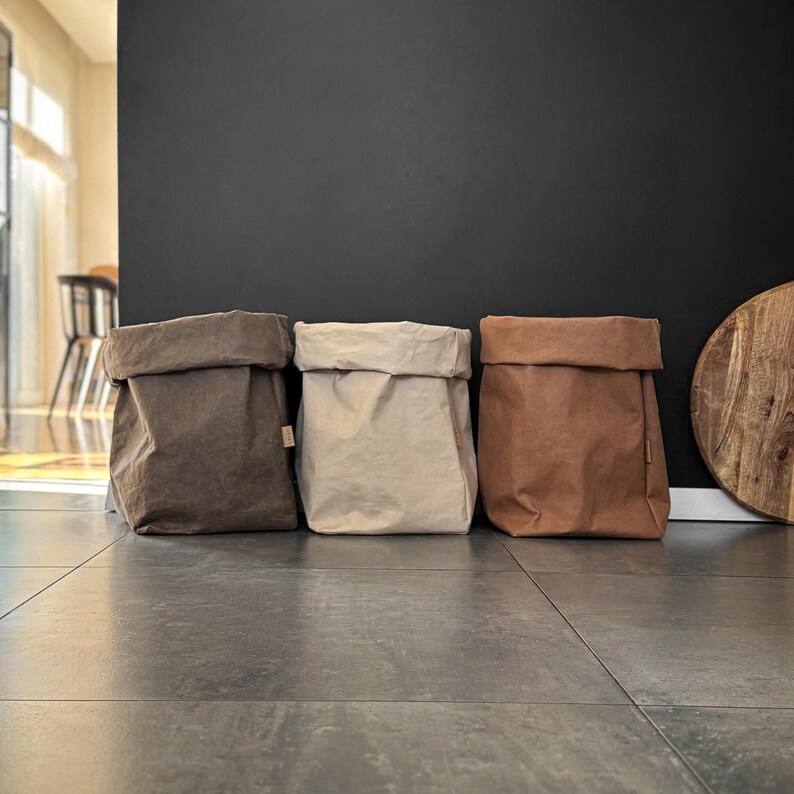 May include: Three rectangular storage bags with folded top edges, arranged in a row. The bags are made of a textured material in shades of dark gray, light beige, and warm tan, each featuring a small rectangular tag on the side seam.