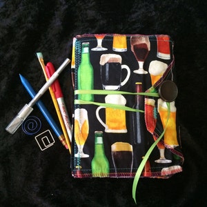 May include: A black fabric journal with a colorful print of beer mugs and bottles. The journal has a green ribbon tied around it and a brown button closure.