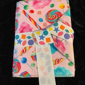 Puede incluir: Un cuaderno de tela rosa con temática de dulces. El cuaderno está envuelto con una cinta blanca con estrellas y formas geométricas de colores. El cuaderno tiene un diseño de dulces coloridos con piruletas, bastones de caramelo y algodón de azúcar.