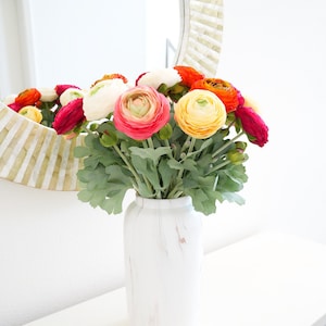 May include: A white vase with a faux marble effect displays a vibrant bouquet of ranunculus flowers. The blooms are in shades of pink, orange, yellow, and red, with green leaves. The vase is on a white table, with a decorative mirror in the background.