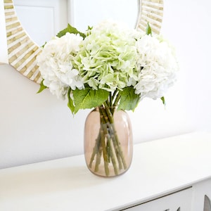 May include: A bouquet of white and green hydrangeas in a clear glass vase with a brown tint. The vase is sitting on a white table.