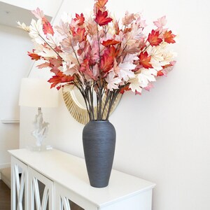 May include: A dark gray ceramic vase with a textured surface holds a bouquet of artificial fall leaves in shades of red, pink, and white. The vase is sitting on a white cabinet with mirrored doors.