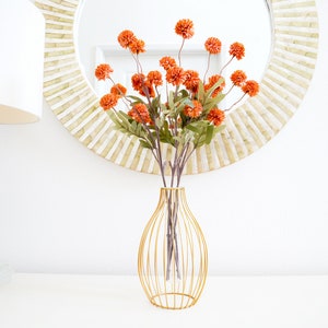 May include: A bouquet of orange silk flowers in a gold wire vase. The vase is on a white surface in front of a round mirror with a white frame.