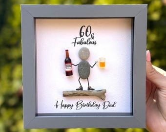 Op de afbeelding: Een vierkant grijs ingelijst wandkunstwerk met een kiezelstenen figuur die op een stuk drijfhout staat. Het figuur houdt een miniatuur bierflesje en een bierpul vast. De tekst luidt 60 & Fabulous en Happy Birthday Dad in zwarte letters.