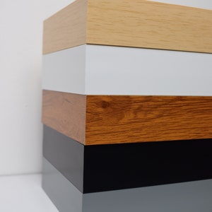 May include: A stack of five wooden picture frames in different colors. The top frame is light wood, followed by white, brown, black, and gray.