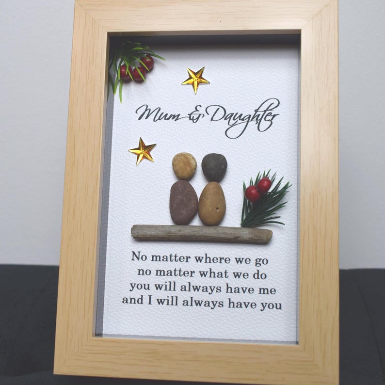 May include: A framed artwork featuring a pebble art design of two figures, representing a mother and daughter, with the text "Mum & Daughter" and a poem about their bond. The artwork is set against a white background with gold stars and a sprig of greenery with red berries.