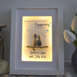 May include: A framed engagement art piece in a white frame. The art features two pebble figures embracing, with a ring detail. The text reads "Congratulations On Your Engagement" and the names "Andrew & Lizzie" with the date "30th July 2026".