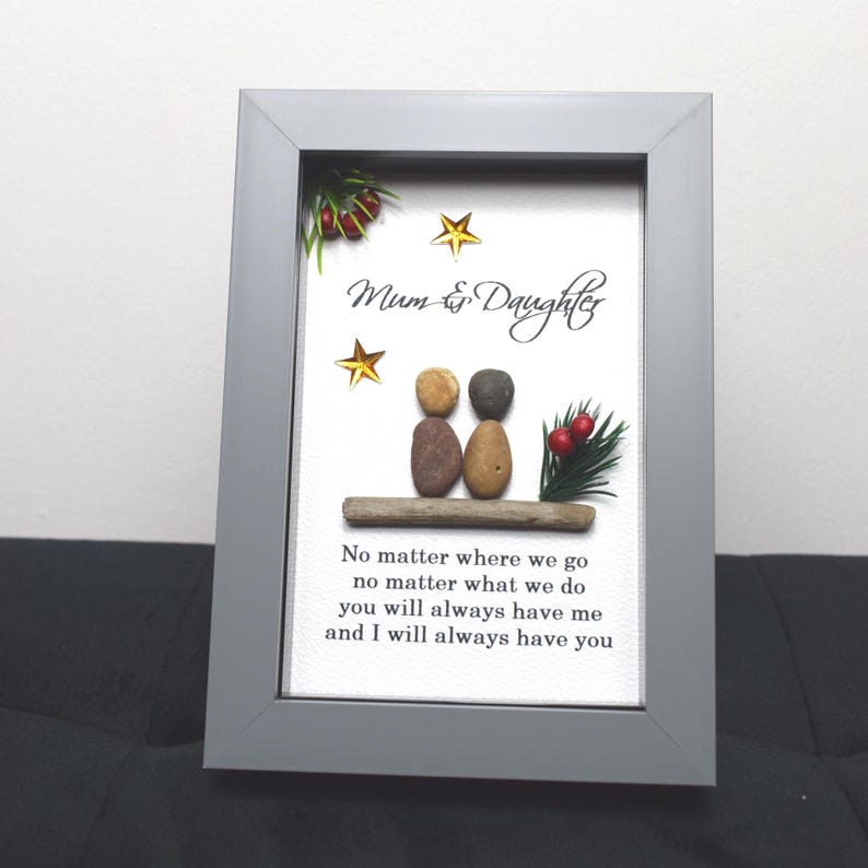May include: A framed pebble art piece with a gray frame and a white background. The art features two figures made of pebbles, a small evergreen sprig with red berries, and gold stars. The text reads "Mum & Daughter" and "No matter where we go, no matter what we do, you will always have me and I will always have you."