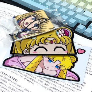 May include: Three colorful car air fresheners featuring a cartoon anime character with blonde hair and a yellow crown. The character is wearing a pink and white dress. The text on the air fresheners reads "来ます" and "帰ります".