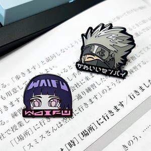 May include: Two enamel pins featuring anime characters. The first pin is a purple and pink anime character with the word "WAIFU" written in white letters. The second pin is a gray and black anime character with the Japanese text "Kawaii Sekai" written in black letters.