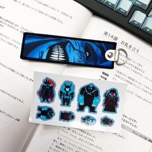May include: A blue and black keychain with a graphic design, and a set of stickers featuring various monster illustrations. The items are placed on top of an open book with Japanese text and a keyboard.
