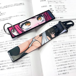 May include: Two colorful anime-style keychains. One keychain features a pink-haired woman with red eyes and a pink and white background. The other keychain features a brown-haired woman with blue eyes and a white background. Both keychains have black straps.
