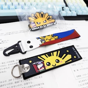 May include: A collection of Filipino-themed accessories, including a keychain, a lanyard, and a pin. The keychain and lanyard feature a smiling sun design with the text "BAHALA KA!" and "AHALA KA SA BUHAY MO". The lanyard has a white strap with black hardware.