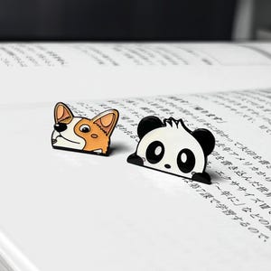May include: Two enamel pins, one featuring a black and white panda peeking over the edge of a page and the other featuring a brown and white corgi peeking over the edge of a page.