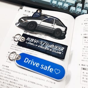 May include: An assortment of automotive-themed accessories, including a black and white car-shaped acrylic charm, a black and blue Lonely Driver's Club keychain, and a blue Drive Safe keychain with a heart symbol.